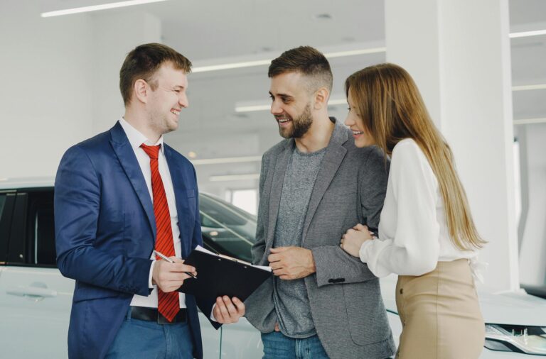 A smiling couple buys a new car from a confident salesman inside a modern car dealership.